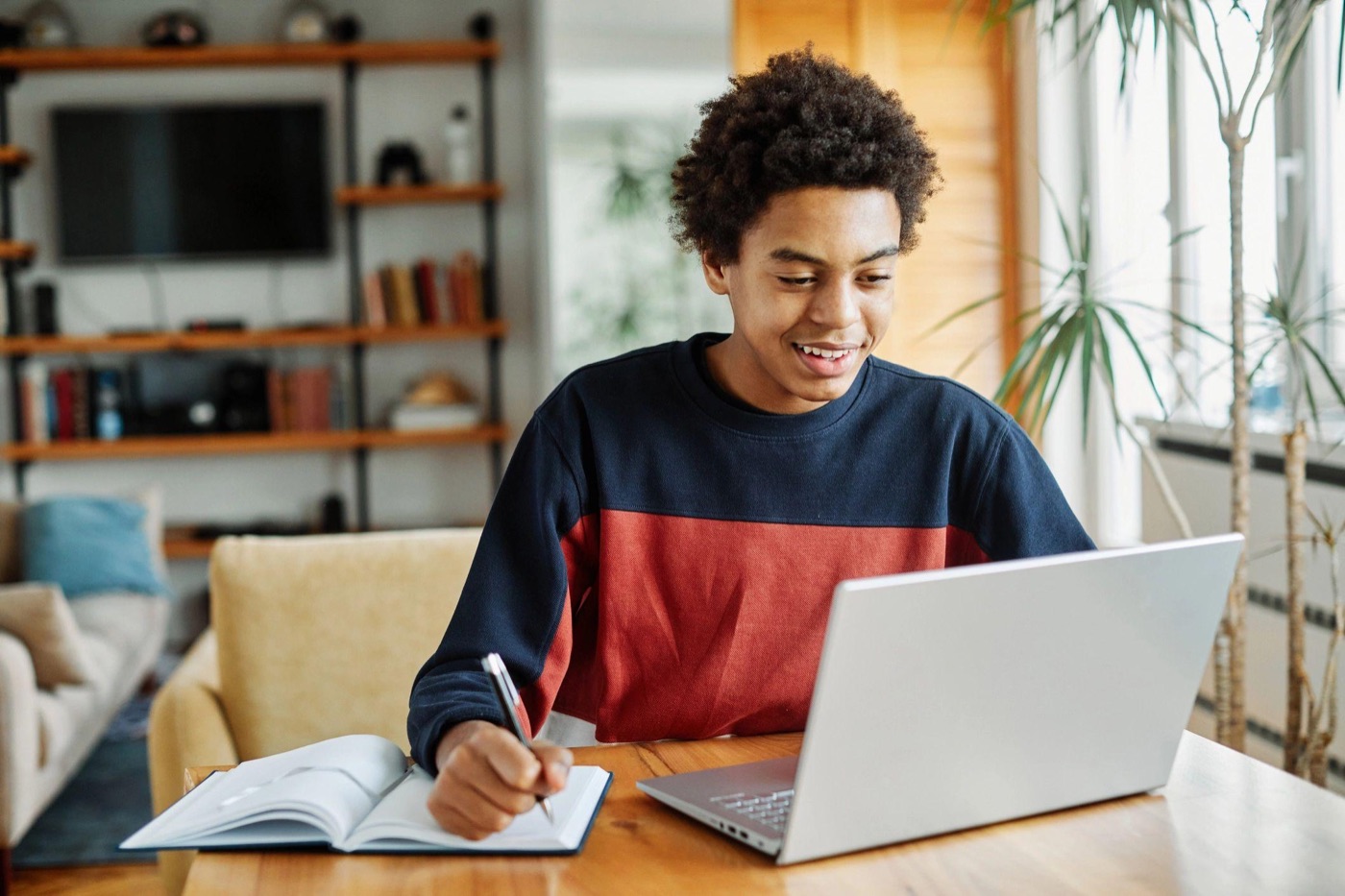 A teenager smiling while writing notes beside a laptop